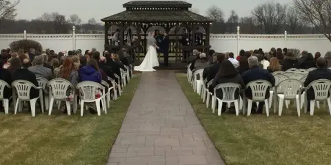 Outdoor wedding ceremony at a gazebo with a paved aisle and seated guests in a landscaped courtyard.