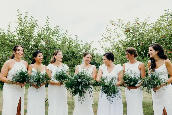 A group of women in white dresses standing outdoors holding large, leafy green wedding bouquets.