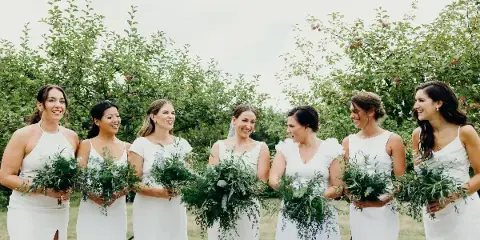 A group of women in white dresses standing outdoors holding large, leafy green wedding bouquets.