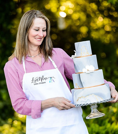 A woman in a LisaMae Bake Shoppe apron holds a four-tier light blue wedding cake with gold accents outdoors.