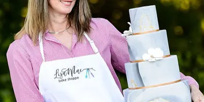 A woman in a LisaMae Bake Shoppe apron holds a four-tier light blue wedding cake with gold accents outdoors.