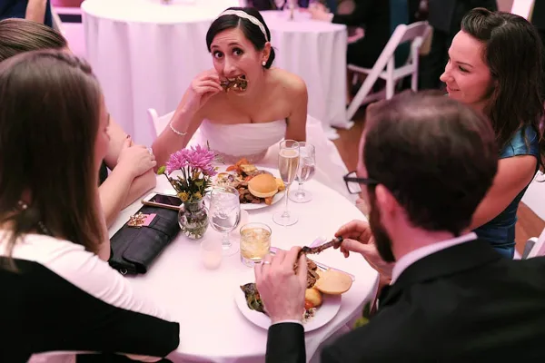 A bride in a white dress eating food at a wedding reception table surrounded by guests.