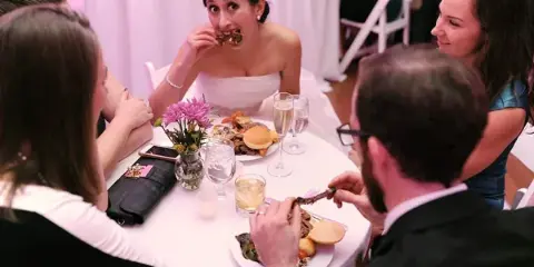 A bride in a white dress eating food at a wedding reception table surrounded by guests.