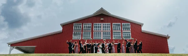A wedding party posing in front of a large red barn with white trim and windows.