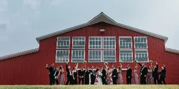 A wedding party posing in front of a large red barn with white trim and windows.