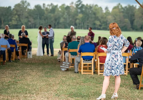 A wedding coordinator holding a clipboard oversees an outdoor ceremony with a bride and groom at the altar and seated guests.