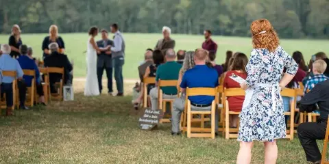 A wedding coordinator holding a clipboard oversees an outdoor ceremony with a bride and groom at the altar and seated guests.