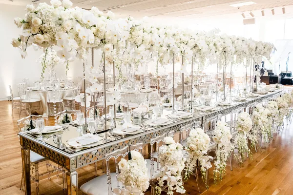 A long, mirrored banquet table set for a formal wedding reception, featuring elaborate white floral arrangements, crystal glassware, and clear acrylic chairs in a bright, modern venue.