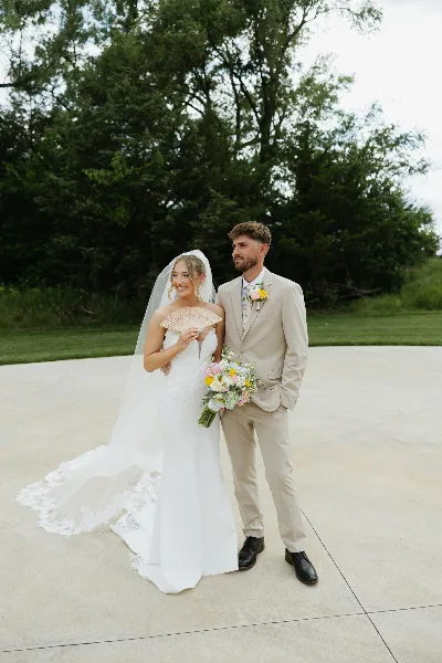 A bride and groom standing together outdoors, the bride holding a fan and a floral bouquet.