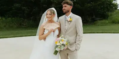 A bride and groom standing together outdoors, the bride holding a fan and a floral bouquet.
