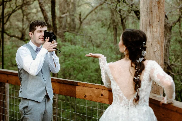 A videographer filming a bride in a white lace wedding dress at an outdoor wooden venue.