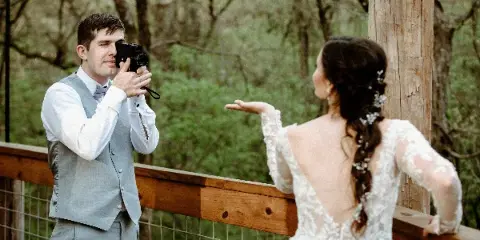 A videographer filming a bride in a white lace wedding dress at an outdoor wooden venue.