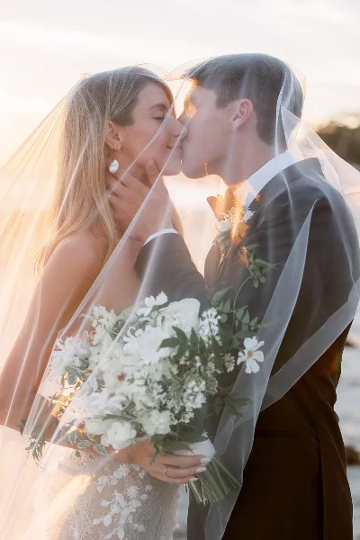 A bride and groom sharing a kiss under a veil with a white floral bouquet in the foreground.