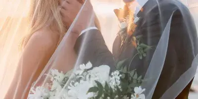 A bride and groom sharing a kiss under a veil with a white floral bouquet in the foreground.