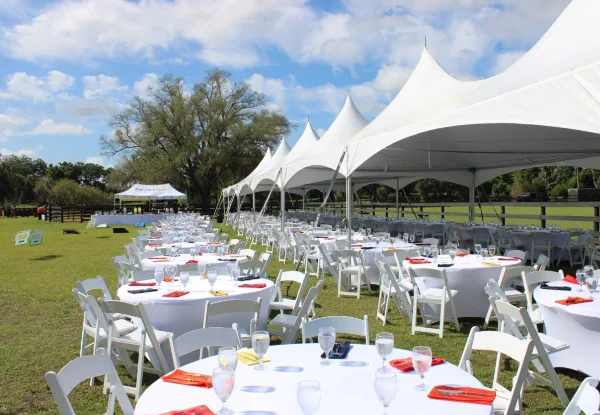 An outdoor event setup featuring a large white peak tent, round tables with white tablecloths, and white chairs arranged on a grassy field.