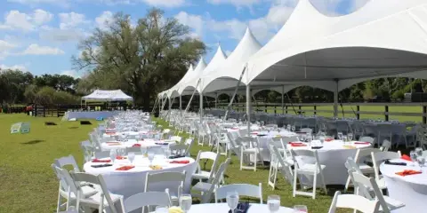 An outdoor event setup featuring a large white peak tent, round tables with white tablecloths, and white chairs arranged on a grassy field.