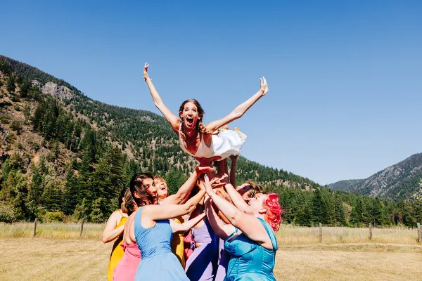 A bride in a white dress is joyfully tossed into the air by a group of smiling bridesmaids in colorful dresses, set against a backdrop of green mountains and a blue sky.