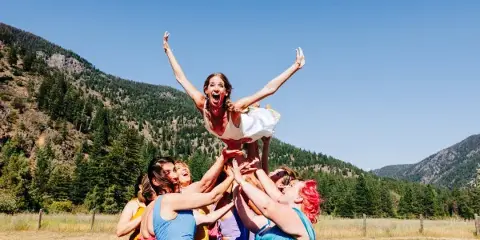 A bride in a white dress is joyfully tossed into the air by a group of smiling bridesmaids in colorful dresses, set against a backdrop of green mountains and a blue sky.