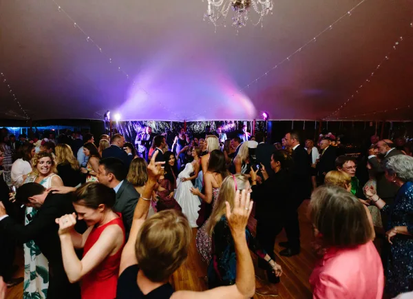 A crowded wedding dance floor with guests dancing under purple stage lighting in a tented venue.
