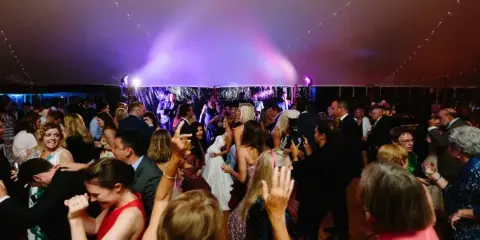 A crowded wedding dance floor with guests dancing under purple stage lighting in a tented venue.