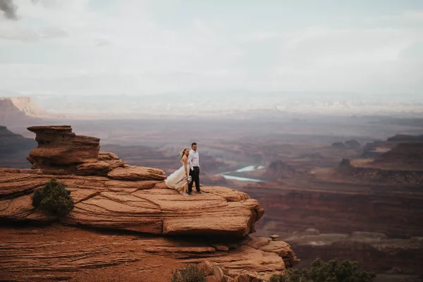 A bride and groom standing on a red rock cliff overlooking a vast canyon landscape.