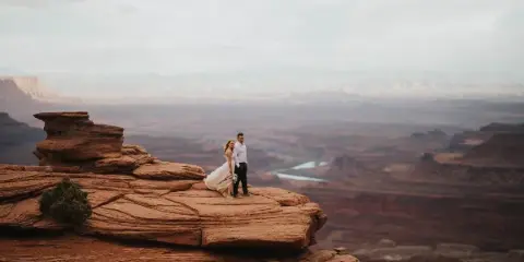 A bride and groom standing on a red rock cliff overlooking a vast canyon landscape.