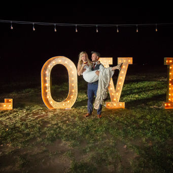 A couple posing with large, illuminated marquee letters spelling 'LOVE' at an outdoor night event.