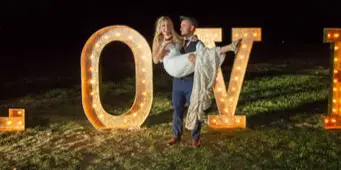 A couple posing with large, illuminated marquee letters spelling 'LOVE' at an outdoor night event.