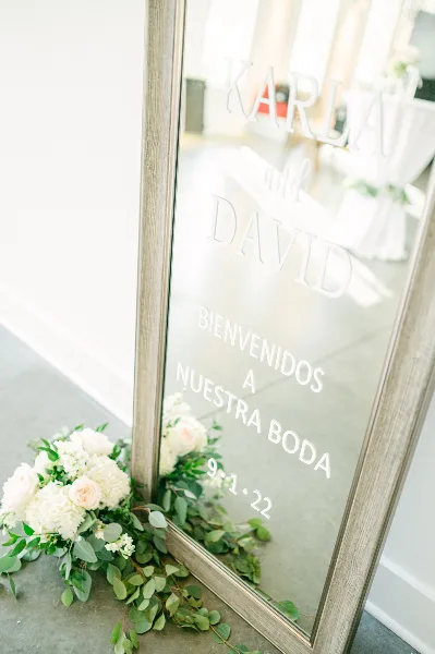 A framed glass wedding welcome sign reading 'Karla and David' with a floral arrangement at the base.