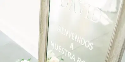 A framed glass wedding welcome sign reading 'Karla and David' with a floral arrangement at the base.
