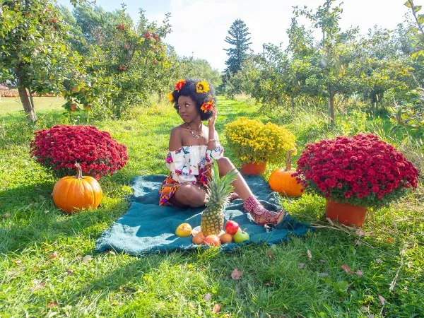 A professional outdoor portrait of a woman sitting on a blue blanket in a grassy field, surrounded by pumpkins, chrysanthemums, and fruit.