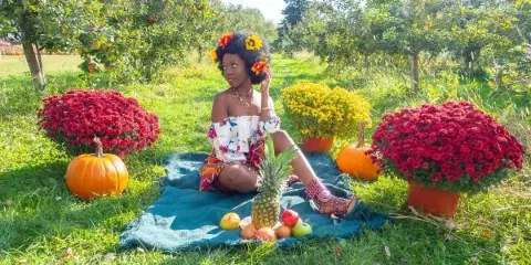 A professional outdoor portrait of a woman sitting on a blue blanket in a grassy field, surrounded by pumpkins, chrysanthemums, and fruit.