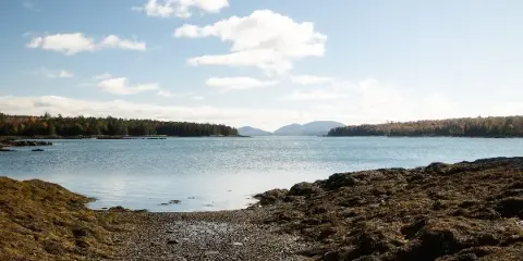 A scenic view of a rocky coastline meeting calm blue water with forested islands in the distance under a partly cloudy sky.