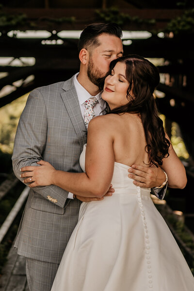 A bride and groom embracing in a romantic pose, with the groom in a grey suit and the bride in a white wedding dress.
