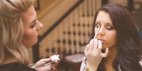 A makeup artist applying makeup to a seated client in an indoor setting.