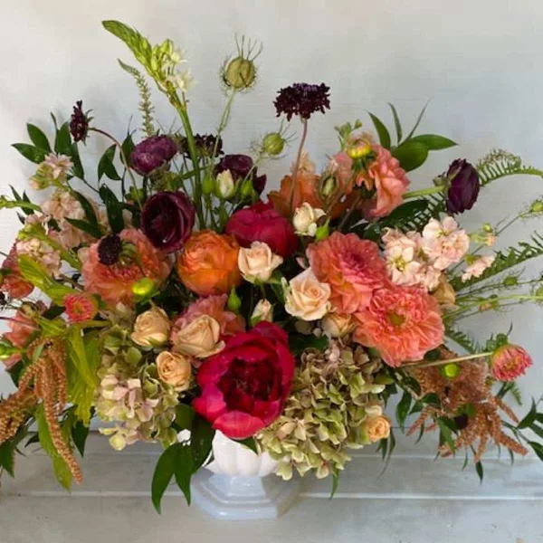 A large, mixed floral arrangement featuring pink, peach, and burgundy blooms in a white vase.