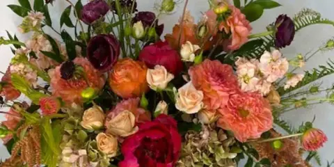 A large, mixed floral arrangement featuring pink, peach, and burgundy blooms in a white vase.