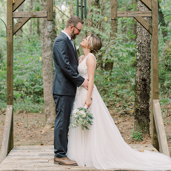 A bride and groom standing together under a wooden arbor in a forest setting.