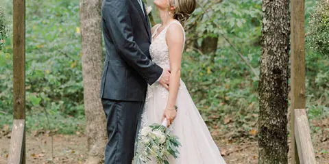 A bride and groom standing together under a wooden arbor in a forest setting.