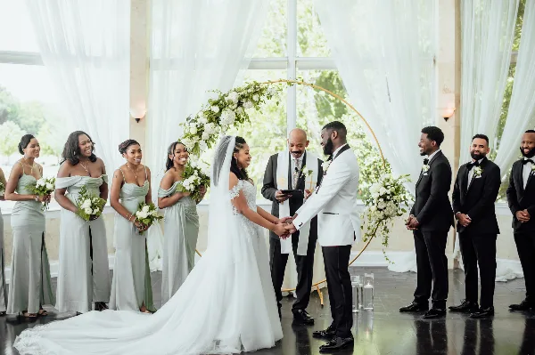A wedding ceremony with a bride and groom exchanging vows under a floral arch, surrounded by bridesmaids and groomsmen.