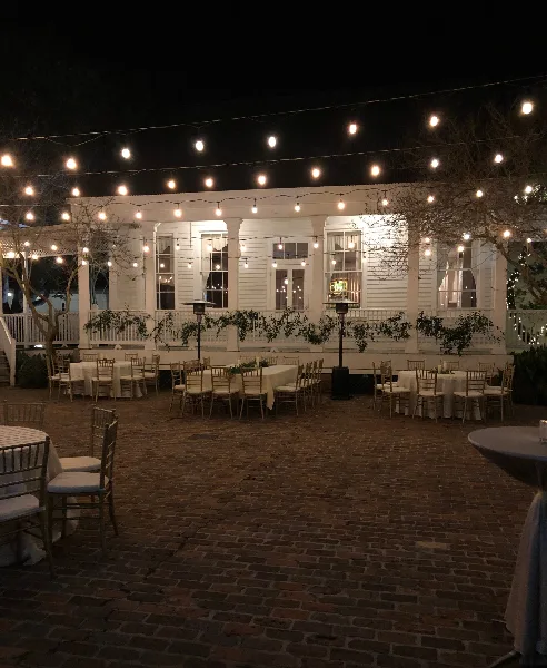 Outdoor brick courtyard at night with white tables, chairs, and string lights in front of a white historic building