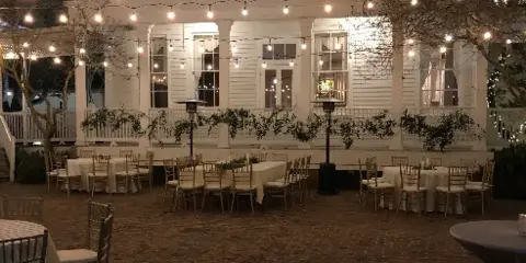 Outdoor brick courtyard at night with white tables, chairs, and string lights in front of a white historic building