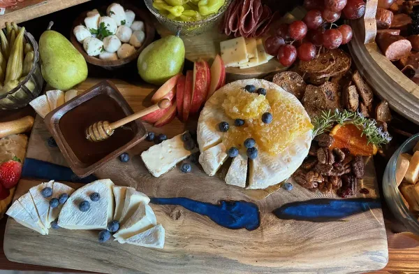 A close-up of a catering spread featuring a large wheel of soft cheese topped with honeycomb and blueberries, accompanied by sliced apples, pears, cured meats, and artisanal bread on a wooden board.