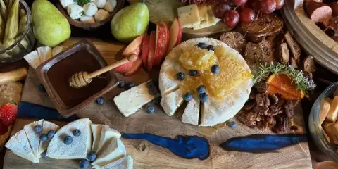A close-up of a catering spread featuring a large wheel of soft cheese topped with honeycomb and blueberries, accompanied by sliced apples, pears, cured meats, and artisanal bread on a wooden board.