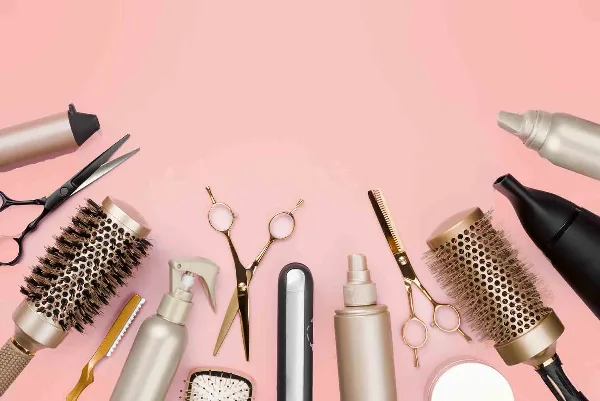 A flat lay arrangement of professional hair styling tools including scissors, brushes, and a flat iron on a pink background.