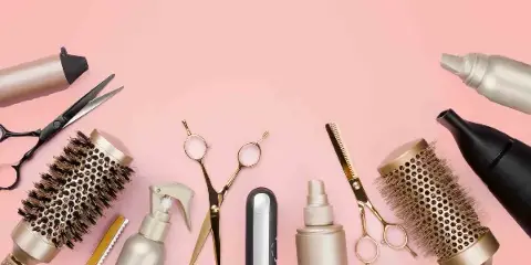 A flat lay arrangement of professional hair styling tools including scissors, brushes, and a flat iron on a pink background.