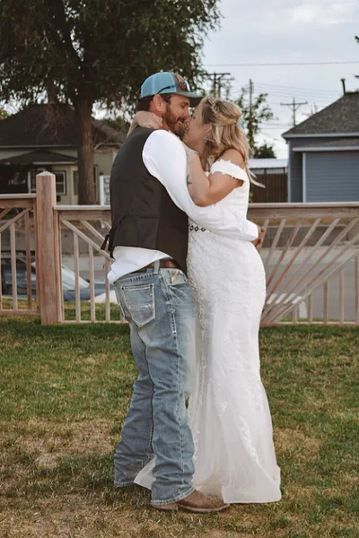 A bride in a white lace wedding dress and a groom in a vest and jeans kissing outdoors on a grassy lawn.
