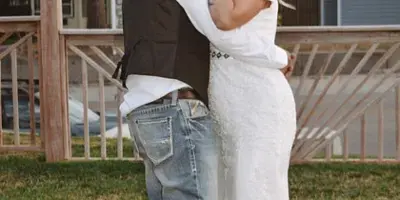 A bride in a white lace wedding dress and a groom in a vest and jeans kissing outdoors on a grassy lawn.