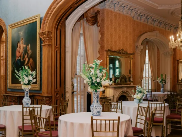An ornate historic room with gold accents and a fireplace, set up with round tables, white tablecloths, and floral arrangements for a wedding or event.