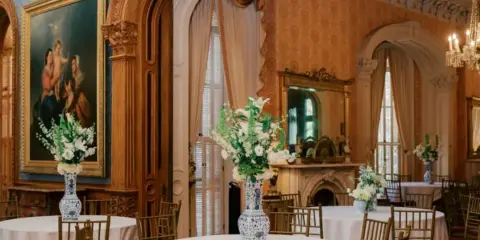 An ornate historic room with gold accents and a fireplace, set up with round tables, white tablecloths, and floral arrangements for a wedding or event.